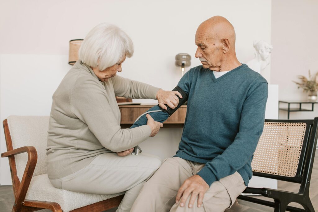 Elderly woman checks man's blood pressure in a cozy living room setting.