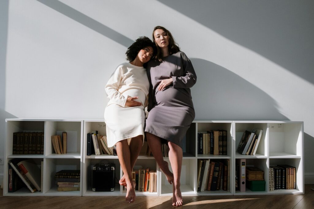 Two pregnant women sitting on a bookshelf, enjoying a peaceful moment together in natural light.