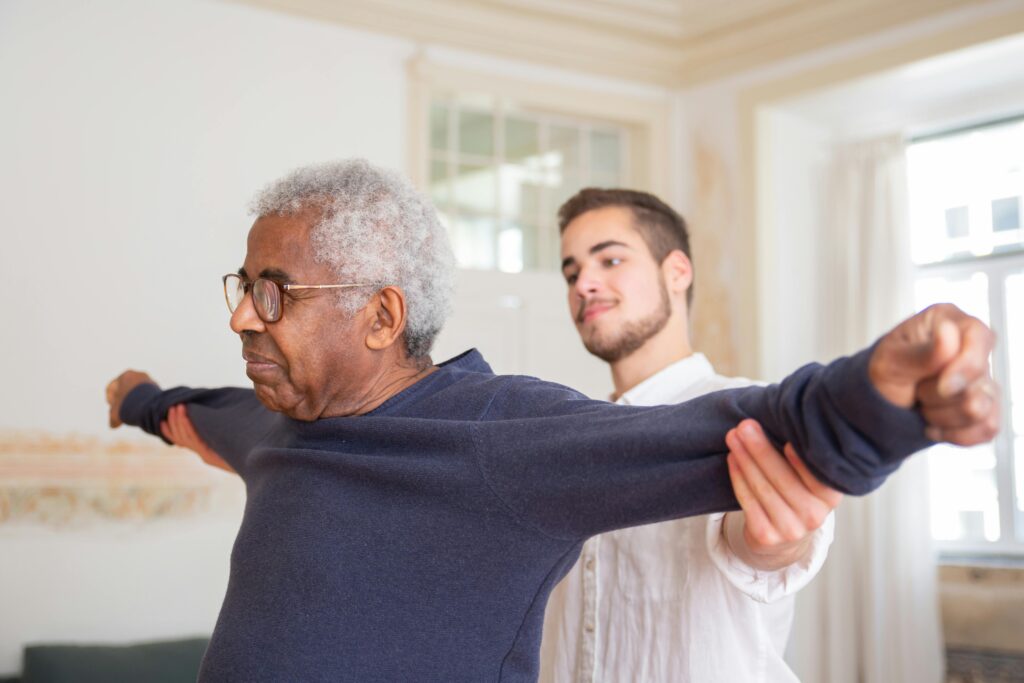 A senior man is assisted by a caregiver for stretching exercises indoors.